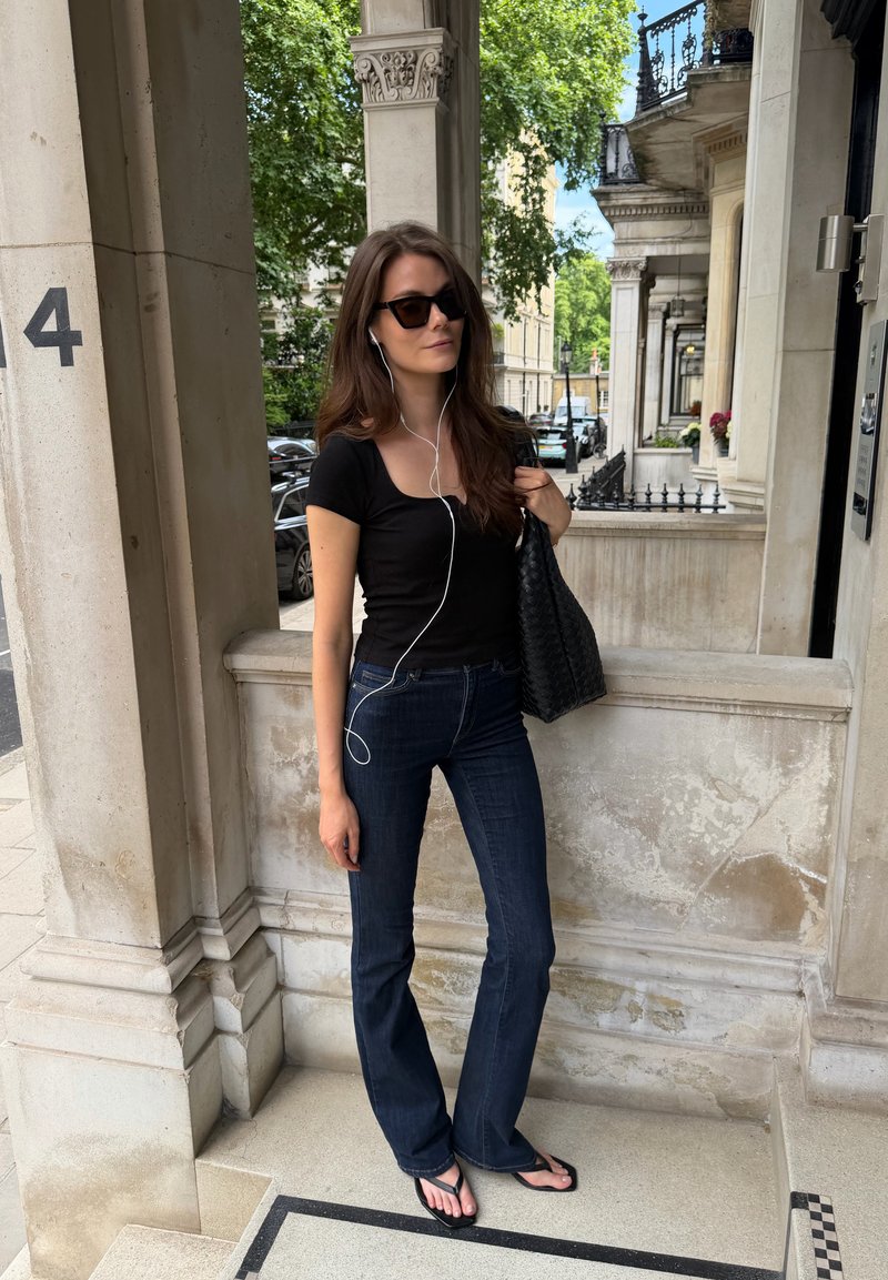 Femme portant des lunettes de soleil, un haut noir, un jean bleu et des sandales, écoutant des écouteurs, tenant un sac noir, debout près d'une colonne en pierre à l'extérieur.