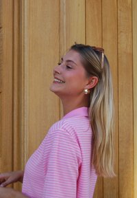 Rosa randig polo med korta ärmar, tillsammans med runda örhängen och solglasögon, mot en träbakgrund.