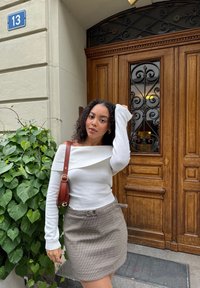 Pull blanc à épaule dénudée et une mini jupe à motif pied-de-poule marron. Un sac marron pend à son épaule, avec des feuillages et une porte en bois en arrière-plan.