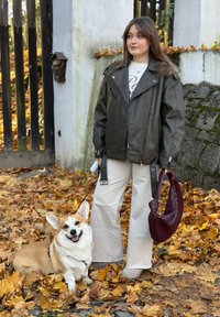 Veste de motard en cuir marron avec quincaillerie argentée, pantalons crème et un sac à main marron foncé. Un Corgi souriant est assis sur des feuilles tombées.