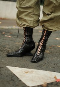 Schwarze Schnürstiefeletten mit Keilabsätzen, getragen zu lockeren olivgrünen Hosen, stehend auf gepflastertem Boden mit einer weißen Pfeilmarkierung.