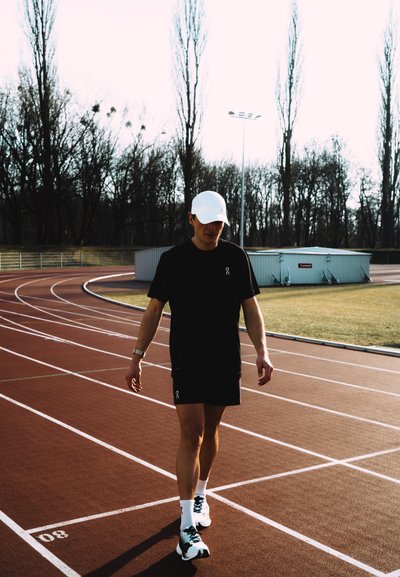 Hombre con ropa deportiva negra y gorra blanca caminando en una pista de atletismo al aire libre cerca de un campo de hierba y árboles sin hojas.