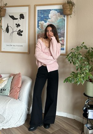 Cardigan de punto rosa, pantalones de pierna ancha negros y botas negras. Ambiente interior con una planta y obras de arte enmarcadas en la pared.