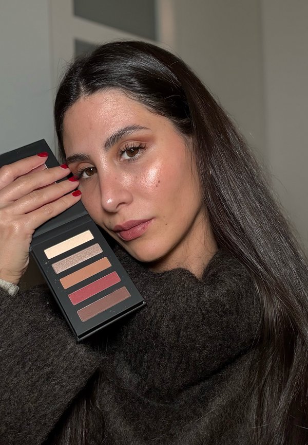 Zeskleuren oogschaduwpalette in een zwarte doos, met matte en glanzende afwerkingen in neutrale tot warme tinten, vastgehouden tegen een donkere trui.