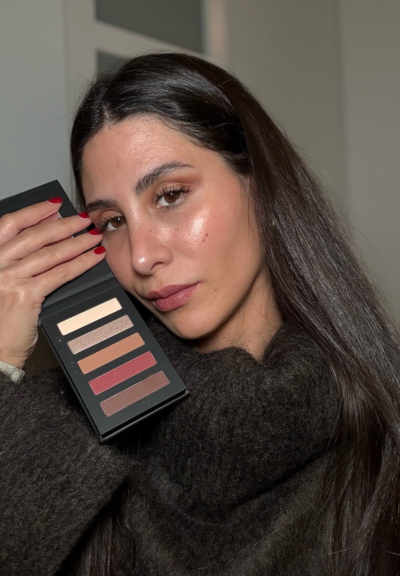 Palette di ombretti a sei colori in una custodia nera, con finiture opache e scintillanti in tonalità neutre e calde, tenuta contro un maglione scuro.