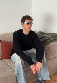 Svart stickad tröja med fransiga muddar, ljusblå slitna jeans, grå sneakers och ljusblå solglasögon på en sittande figur. Varmtonad soffbakgrund.