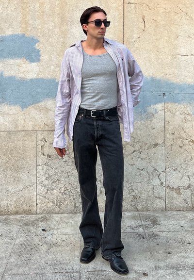 Pantalones de mezclilla negra combinados con una camiseta sin mangas gris claro y una camisa de cuadros de gran tamaño. Los accesorios incluyen gafas de sol negras. Textura suave.