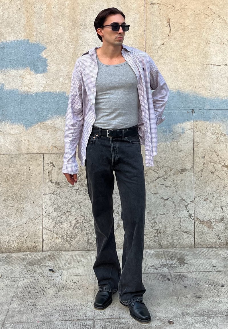 Calças de ganga pretas combinadas com uma camisola sem mangas cinza clara e uma camisa xadrez oversized. Os acessórios incluem óculos de sol pretos. Textura suave.