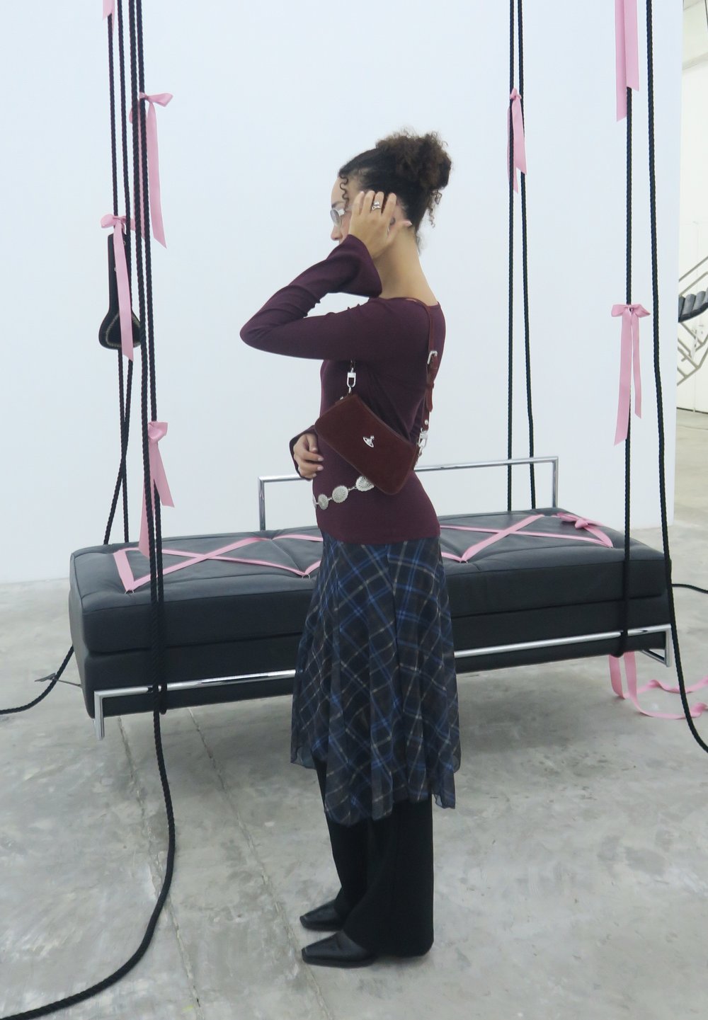 A woman wearing glasses, a burgundy top, and a plaid skirt stands next to a suspended black bench adorned with pink ribbons tied to black ropes.