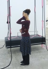 A woman wearing glasses, a burgundy top, and a plaid skirt stands next to a suspended black bench adorned with pink ribbons tied to black ropes.