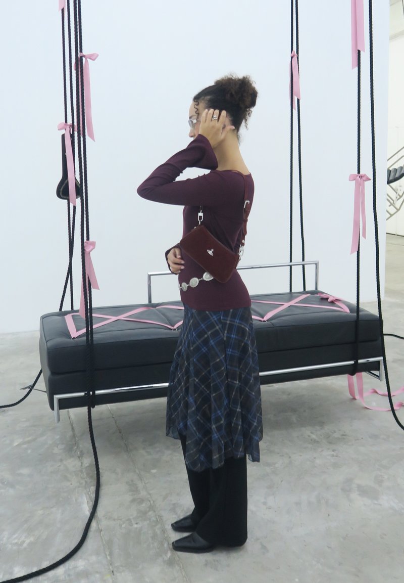 Žena u naočalama, tamnocrvenoj majici i kariranoj suknji stoji pored suspendirane crne klupe s ružičastim trakama vezanim za crne uže.