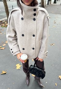 Jasnoszary wełniany płaszcz dwurzędowy z czarnymi guzikami, w połączeniu z ciemnobrązowymi spodniami i szpilkami z zaostrzonymi noskami. Trzyma filiżankę kawy i czarną torebkę.