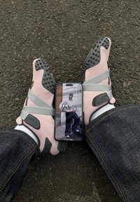 Persoon met roze en grijze Puma-schoenen en spijkerbroek, met een smartphone waarop een man in casual streetwear buiten zit, te zien is.