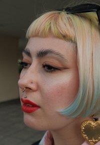 Kort, pastellfarget hår med grønne bånd, gull septum og stiftpiercinger, dristige røde lepper, og en hjerteformet gulløredobb.