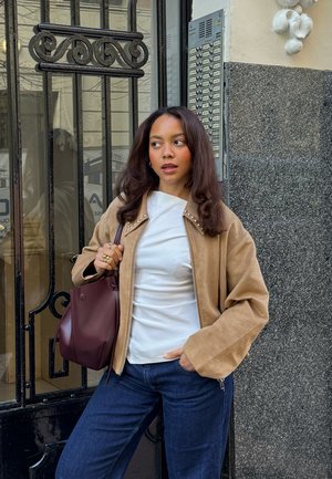 Mujer con cabello castaño que lleva una chaqueta beige, blusa blanca y jeans azules, sosteniendo un bolso granate, de pie junto a una reja metálica negra y la entrada de un edificio.