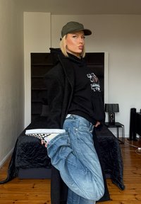 Sweat à capuche noir surdimensionné, manteau foncé, jean bleu et baskets à motif à carreaux. Environnement intérieur avec sol en bois et literie sombre.