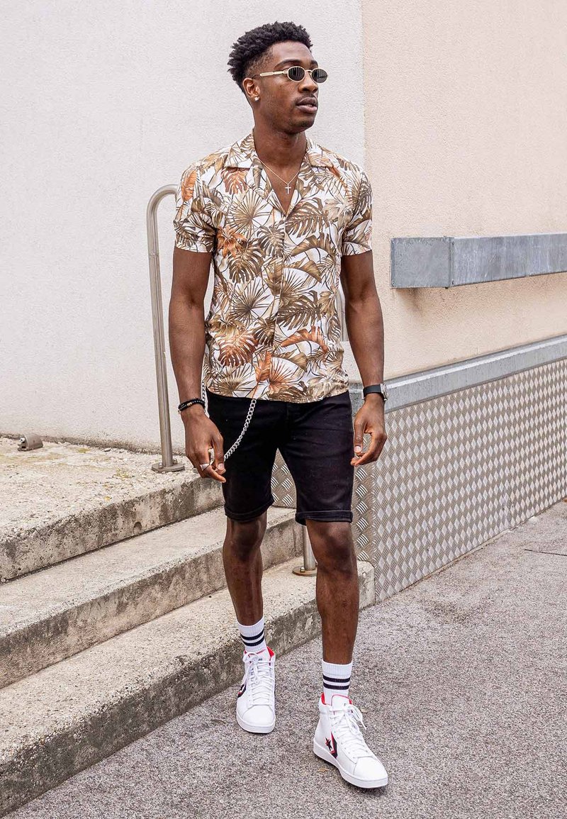 Short-sleeved brown and cream floral shirt, black shorts, white high-top trainers, and sunglasses, accessorised with a silver chain.