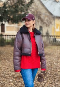 Jachetă maro scurtă din piele cu guler de blană artificială neagră, pulover tricotat roșu, blugi albaștri și o geantă mică maro din blană, pe fundal exterior.