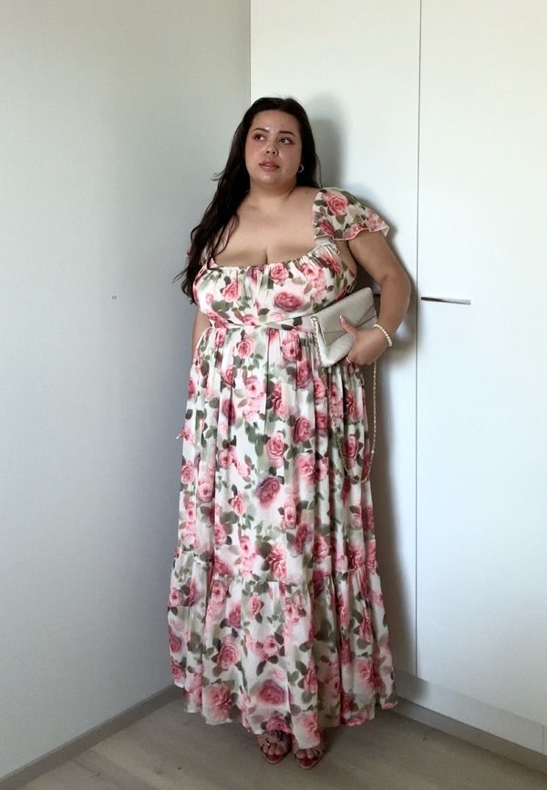 Eine Frau in einem langen, rosa geblümten Kleid steht drinnen, hält eine beige Clutch, mit offenem Haar und neutralem Gesichtsausdruck, neben einem weißen Kleiderschrank.