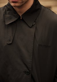 Must mustveekattega kõrge krae, esiküljel nööpkinnitus ja tikitud logo "WRSTBHVR". Sile, matt tekstuur.