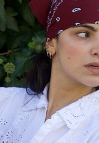 Femme aux cheveux foncés portant un bandana rouge à motifs, des boucles d'oreilles créoles en or et un chemisier blanc en broderie anglaise, regardant sur le côté avec un arrière-plan végétal.