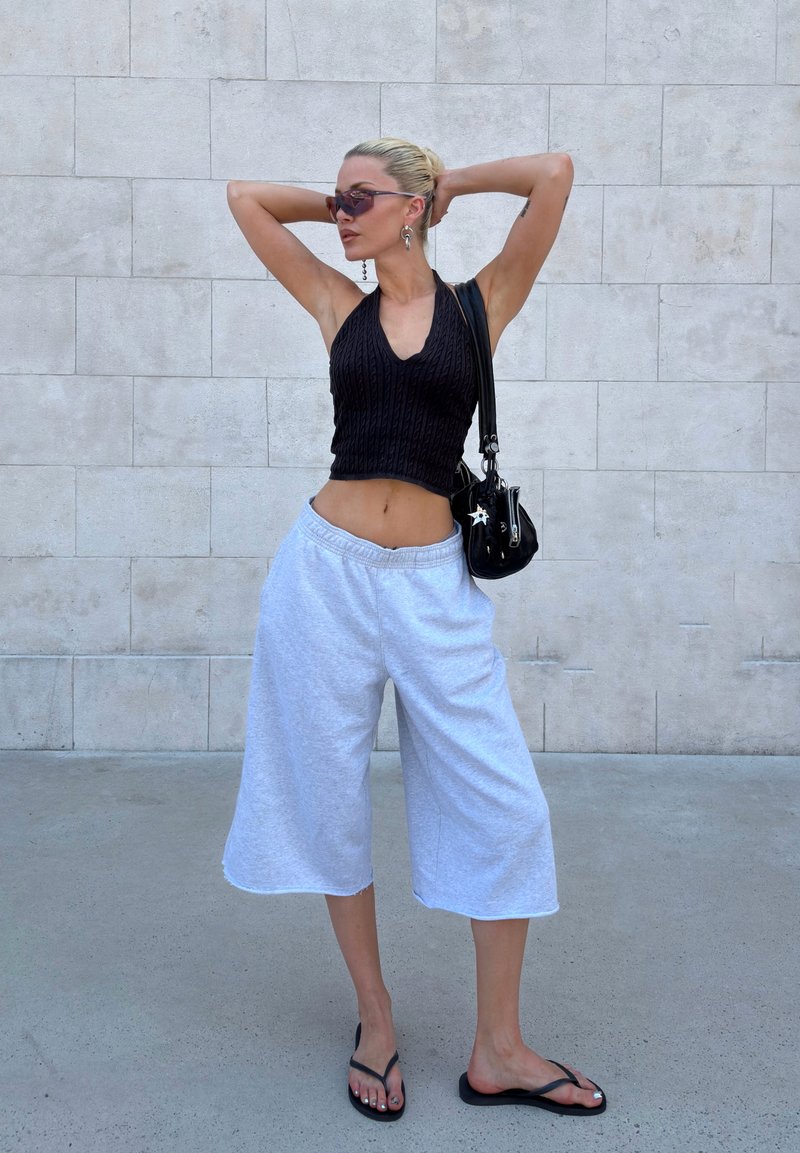 Femme avec des lunettes de soleil portant un haut noir sans manches, un pantalon blanc à jambes larges et des tongs noires, debout contre un mur carrelé avec les mains derrière la tête.
