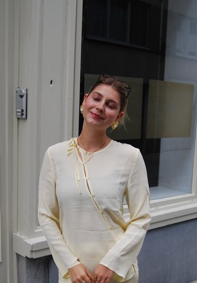 Blusa color crema con detalle de lazo, confeccionada en un tejido ligeramente transparente y de manga larga; complementada con pendientes y collar en forma de corazón.