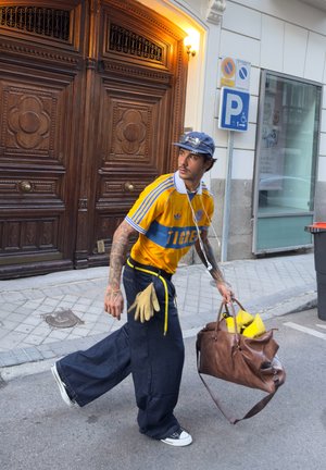 Jersey amarillo con banda azul, pantalones oscuros anchos, bolso de cuero marrón y guantes. Gafas de sol brillantes en la gorra; caminando por una calle de adoquines.