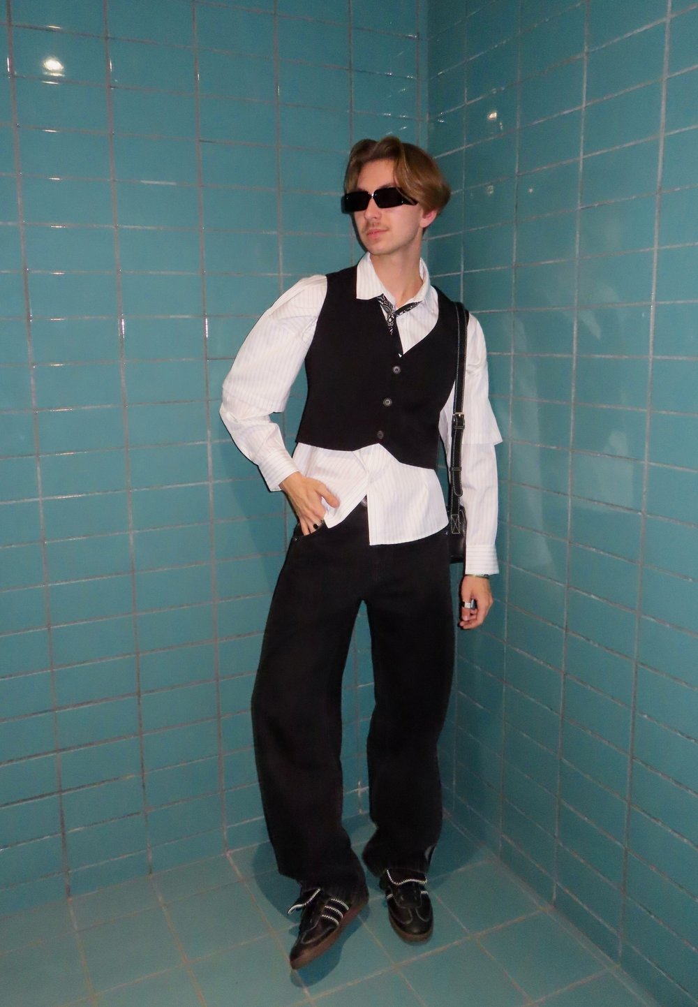 Melns veste pār baltu svītrainu kreklu, melnas platas bikses un saulesbrilles. Uzkrītošs kontrasts ar tirkīza flīzēto fonu.