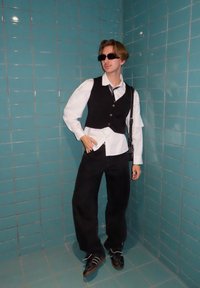 Melns veste pār baltu svītrainu kreklu, melnas platas bikses un saulesbrilles. Uzkrītošs kontrasts ar tirkīza flīzēto fonu.