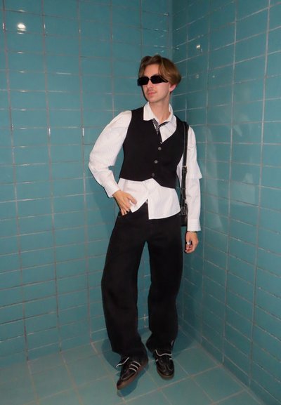 Chaleco negro sobre una camisa blanca con rayas, pantalones negros de pierna ancha y gafas de sol. Contraste notable con el fondo de azulejos turquesa.