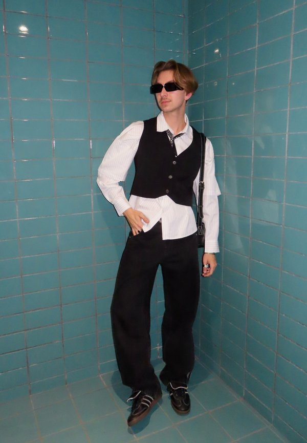 Colete preto por cima de uma camisa branca às riscas, calças pretas de perna larga e óculos de sol. Contraste notável com o fundo de azulejos turquesa.