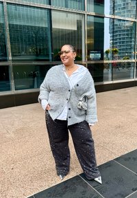 Grijze gebreide cardigan over een wit shirt, gecombineerd met zwarte broek met zilveren studs en puntige zilveren hakken. Kleine zilveren handtas.