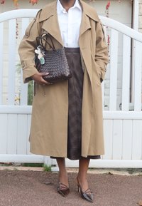 Beżowy płaszcz przeciwdeszczowy na białej koszuli, zestawiony z brązową spódnicą w kratę. Trzyma ciemną plecioną torebkę i nosi brązowe szpilki z ostrym noskiem.