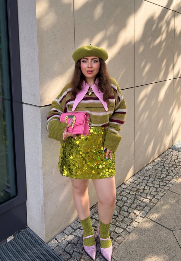 Femme portant un béret vert, un pull rayé, une jupe dorée à sequins, des chaussettes vertes, des chaussures roses et tenant un sac à main rose, debout près d'un mur beige.