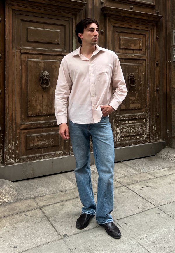 Hombre con camisa rosa claro abotonada, vaqueros azules y zapatos negros, de pie sobre un pavimento de piedra frente a unas grandes puertas dobles de madera envejecida.
