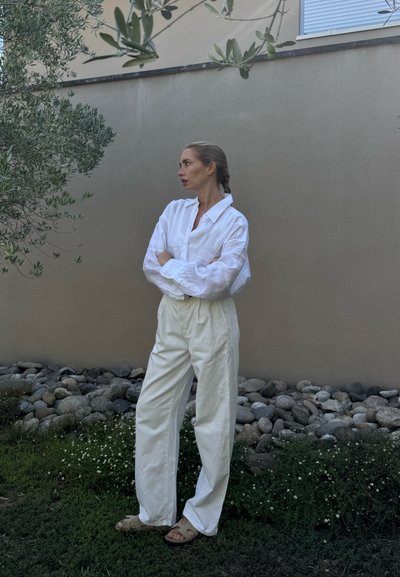 Camisa de botones blanca de lino y pantalones holgados color blanco roto, combinados con sandalias ligeras, frente a una pared neutra y un fondo de piedra natural.