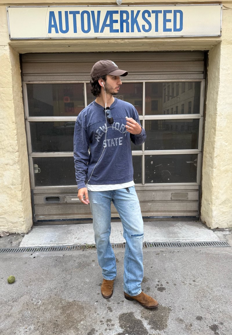 Jeune homme avec casquette, sweatshirt et jean, debout devant une porte de garage avec le panneau « AUTOVÆRKSTED », tenant des écouteurs et regardant de côté.