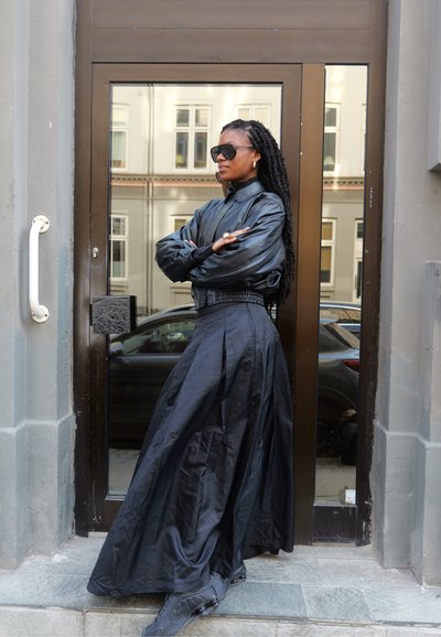 Chaqueta bomber de cuero negra con un cuello alto, combinada con una falda larga y fluida negra. Gafas de sol grandes y elegantes y calzado negro.