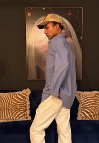 Νέος άνδρας με μπεζ καπέλο και μπλε πουκάμισο περπατάει μέσα σε κλειστό χώρο, με πίνακες έργων τέχνης και έναν σκούρο μπλε καναπέ με μαξιλάρια με σχέδια στο φόντο.