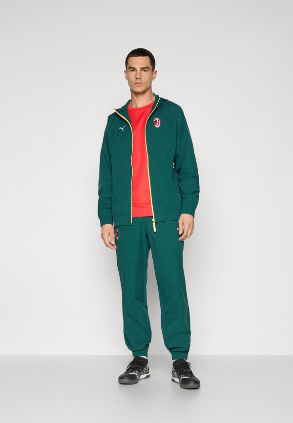 Zielony sportowy dres składający się z kurtki zapinanej na zamek z żółtymi akcentami i logo, zestawiony z pasującymi spodniami. Model ma na sobie czarne buty.