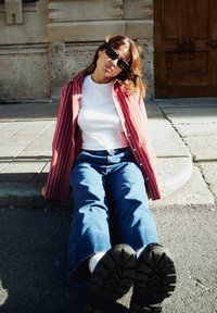 Camicia a righe rosse e bianche sopra una maglietta bianca, abbinata a jeans a gamba larga blu. Occhiali da sole neri e calzature nere testurizzate.