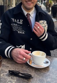 Casaco varsity preto com riscas brancas, logo de Yale, usado sobre uma camisa branca e uma gravata bordô. Segurando um copo branco sobre um prato branco com uma colher.