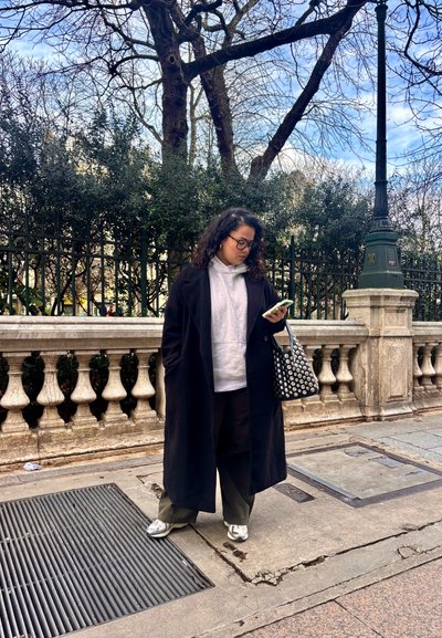 Abrigo largo negro, sudadera gris, pantalones negros holgados, zapatillas plateadas. Bolsa negra con lunares blancos. De pie junto a una barandilla de piedra con árboles.