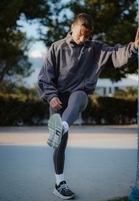 Nő sportos ruházatban nyújtja az egyik lábát egy hálóoszlopon szabadtéren, napk�özben, fákkal körülvéve és elmosódott háttér előtt.