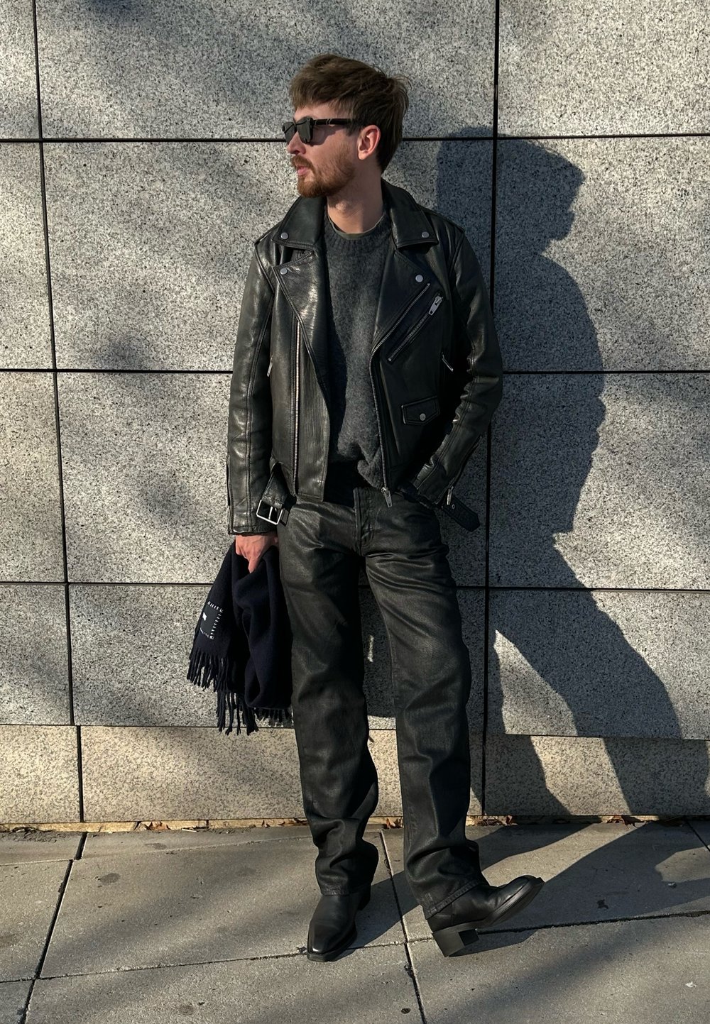 Fekete bőr motorosdzseki szürke pulóver fölött, fekete farmerrel és bokacsizmával párosítva. Egy tengerészkék sál van a karjára terítve.