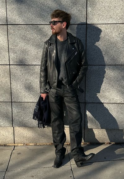 Chaqueta de cuero negra tipo motorista sobre un suéter gris, combinada con jeans negros y botines. Una bufanda azul marino está colgada sobre su brazo.