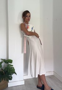 Weißes, geripptes Midikleid mit einem seitlichen Schlitz, kombiniert mit einer mehrfarbigen, floral gemusterten Clutch und dunkelblauen Slippern. Innenraum.