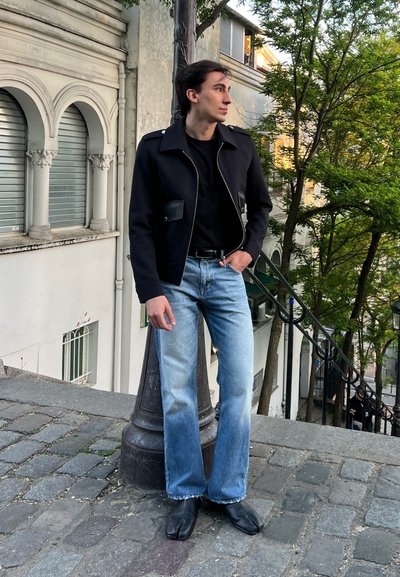 Joven en chaqueta negra y jeans azules está de pie en una calle empedrada junto a un farola, mirando a la derecha con árboles y escaleras detrás de él.