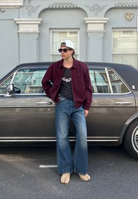 Un giovane indossando un cappellino bianco, occhiali da sole, una giacca bordeaux, una camicia nera, jeans a zampa d'elefante e scarpe beige si trova accanto a una macchina d'epoca nera su una strada.